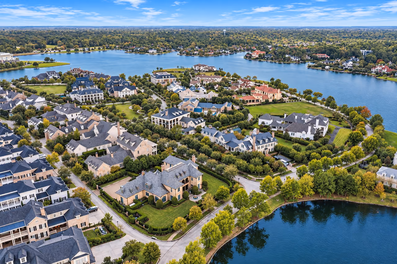 Aerial view of houses beside a wide lake: pale brick and stone houses with steep gray and blue roofs stand among clipped hedges and trees starting to turn yellow and orange. In the foreground a large house sits behind a hedged lawn and a round fountain. Across the lake, more houses line a wooded shore, and a water tower stands on the horizon.